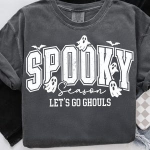 May include: Dark gray t-shirt with the words "Spooky Season" in a white, bold font, along with ghost and bat graphics. The phrase "Let's Go Ghouls" is also printed on the shirt, perfect for Halloween.