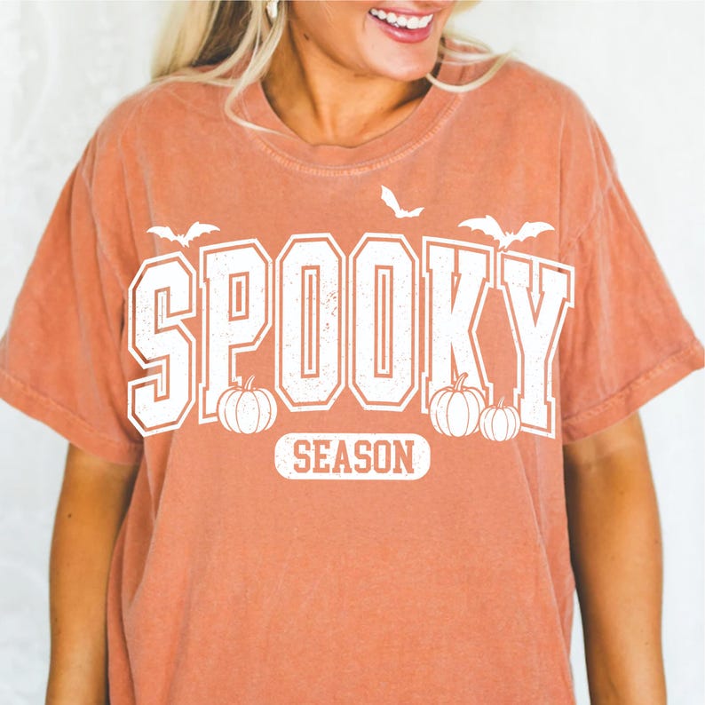 May include: A burnt orange t-shirt with the words "Spooky Season" in white block letters. The design includes white bats and pumpkins, perfect for Halloween. The shirt has a vintage, slightly faded look.