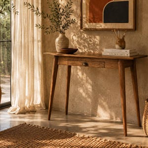 Op de afbeelding: Een houten sidetable met vier taps toelopende poten en een enkele centrale lade met een donkere metalen greep. Op het rechthoekige tafelblad staan een keramische vaas met groen, een kleine donkere kom en twee boeken getiteld The Kinfolk Home en Live Beautiful.