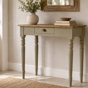 Op de afbeelding: Een saliegroene sidetable met een blad van natuurlijk hout en een enkele centrale lade met een messingkleurige halvemaanvormige greep. De tafel staat op vier gedraaide, gecanneleerde poten en is voorzien van een keramische vaas en boeken.