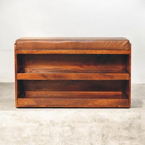 May include: A wooden shoe rack with a brown leather cushioned top and two shelves. The shoe rack is made of dark wood and has a simple design.