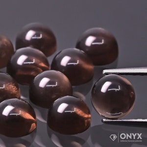 Op de afbeelding: Een groep van 12 ronde, cabochon geslepen rookkwarts edelstenen. De stenen zijn donkerbruin van kleur met een lichte glans. De stenen zijn los op een grijs oppervlak gerangschikt.