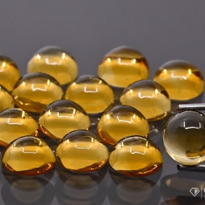 Könnte beinhalten: Eine Gruppe von 15 runden, durchscheinenden, gelb-orangen Cabochon-Edelsteinen, wobei ein Edelstein mit einer Pinzette gehalten wird. Die Edelsteine sind in einem Halbkreis auf einer grauen Oberfläche angeordnet.
