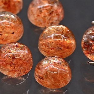 Könnte beinhalten: Eine Sammlung runder, polierter Edelsteine mit einem warmen, rötlich-orangen Farbton. Die Edelsteine haben ein durchscheinendes Aussehen mit inneren Mustern, und einer wird von einer Pinzette gehalten. Die Edelsteine sind wahrscheinlich Sonnenstein oder ähnliches.