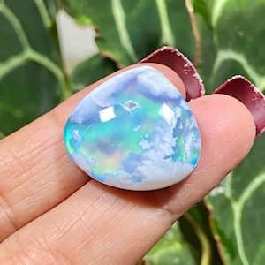 May include: A pear-shaped opal gemstone with a cloudy, iridescent appearance. The stone displays a mix of blue, green, and white hues, with a subtle play of light. The gemstone is held in a hand, showcasing its polished surface.