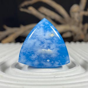 Puede incluir: Una gema triangular pulida, de un vibrante color azul, que recuerda a un cielo despejado. La piedra presenta inclusiones blancas en forma de nube, creando un patrón único. Descansa sobre una base circular blanca, mostrando su forma y color.