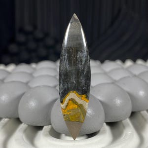 Op de afbeelding: Een gepolijste, langwerpige edelsteen met een puntige punt. De steen heeft een kleurengradiënt, waaronder helder, grijs, zwart, wit en geel. Het onderste deel heeft een bruine punt. De edelsteen wordt op een grijze ondergrond getoond.