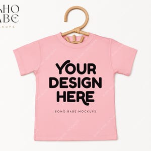 May include: Pink t-shirt mockup with a hanger. The shirt has the text "YOUR DESIGN HERE" and "BOHO BABE MOCKUPS" printed on it.