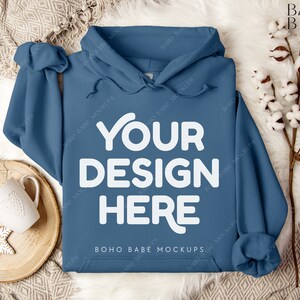 Puede incluir: Una sudadera con capucha azul con el texto "YOUR DESIGN HERE" en blanco en la parte delantera. La sudadera est&aacute; sobre una superficie blanca y esponjosa con una bandeja de madera y una taza blanca.