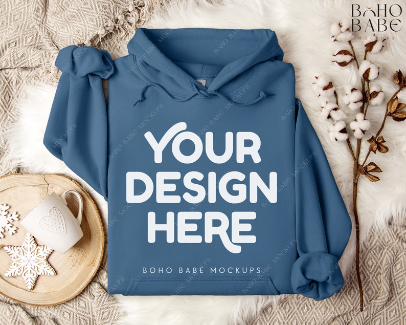 Puede incluir: Una sudadera con capucha azul con el texto "YOUR DESIGN HERE" en blanco en la parte delantera. La sudadera est&aacute; sobre una superficie blanca y esponjosa con una bandeja de madera y una taza blanca.