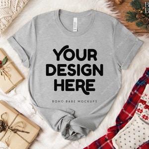 May include: A heather gray t-shirt with the text "YOUR DESIGN HERE" in bold black letters. The shirt is laid on a white fluffy surface, with wrapped gifts and holiday-themed socks nearby. The words "BOHO BABE MOCKUPS" are at the bottom.