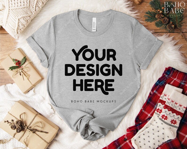 May include: A heather gray t-shirt with the text "YOUR DESIGN HERE" in bold black letters. The shirt is laid on a white fluffy surface, with wrapped gifts and holiday-themed socks nearby. The words "BOHO BABE MOCKUPS" are at the bottom.