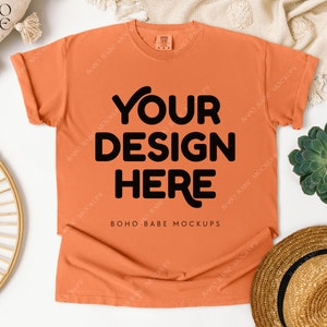May include: An orange t-shirt with the text "YOUR DESIGN HERE" in black. The shirt is laid flat on a white surface, with a straw hat, succulent, and decorative elements around it. The shirt is a mock-up.