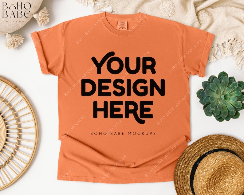May include: An orange t-shirt with the text "YOUR DESIGN HERE" in black. The shirt is laid flat on a white surface, with a straw hat, succulent, and decorative elements around it. The shirt is a mock-up.