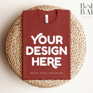 Könnte beinhalten: Ein rotes T-Shirt mit dem Text "YOUR DESIGN HERE" in Weiß auf einer geflochtenen Strohmatte. Der Text "BOHO BABE MOCKUPS" ist ebenfalls auf dem T-Shirt zu sehen.