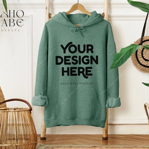 May include: A green hoodie with the text "YOUR DESIGN HERE" in black, hanging on a wooden hanger. The sleeves are cuffed. The background includes a wooden ladder, a woven basket, and a round woven bag.