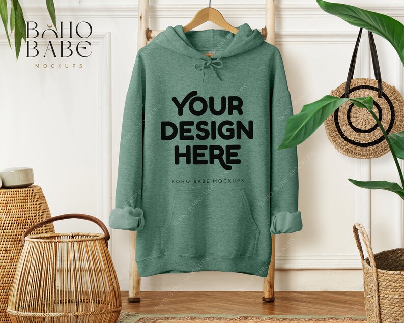 May include: A green hoodie with the text "YOUR DESIGN HERE" in black, hanging on a wooden hanger. The sleeves are cuffed. The background includes a wooden ladder, a woven basket, and a round woven bag.