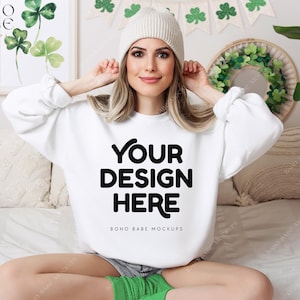 May include: White sweatshirt with the text "Your Design Here" and "Boho Babe Mockups" printed on the front. The model is wearing a white beanie and sitting on a white bed.