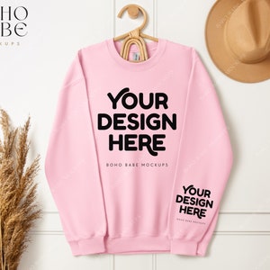 May include: A pink crewneck sweatshirt mockup with the text "YOUR DESIGN HERE" on the front and back. The sweatshirt is hanging on a hanger against a white wall.