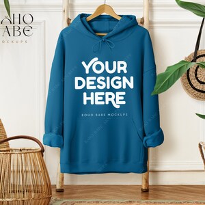 May include: Teal hoodie with the text "YOUR DESIGN HERE" in white, hanging on a wooden ladder. The hoodie has a drawstring hood and a front pocket. The image includes a woven basket, a round woven bag, and greenery.