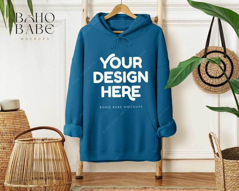May include: Teal hoodie with the text "YOUR DESIGN HERE" in white, hanging on a wooden ladder. The hoodie has a drawstring hood and a front pocket. The image includes a woven basket, a round woven bag, and greenery.