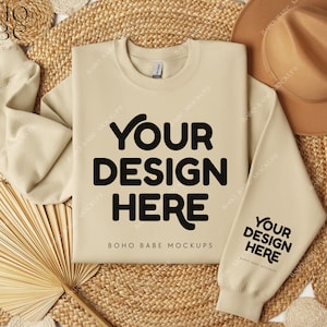 May include: A beige sweatshirt with the text "YOUR DESIGN HERE" on the front and "YOUR DESIGN HERE" on the sleeve. The sweatshirt is laid flat on a woven straw mat with a brown hat and a dried palm leaf fan.