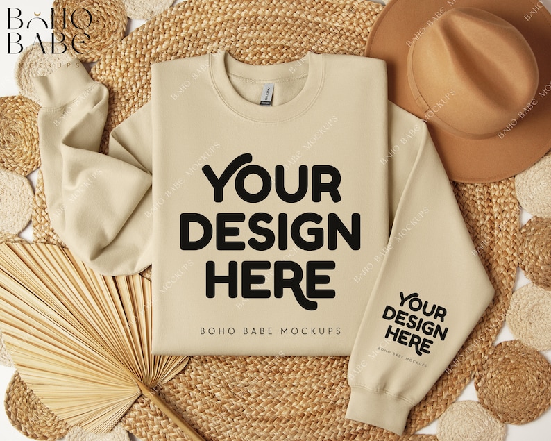 May include: A beige sweatshirt with the text "YOUR DESIGN HERE" on the front and "YOUR DESIGN HERE" on the sleeve. The sweatshirt is laid flat on a woven straw mat with a brown hat and a dried palm leaf fan.