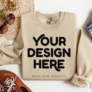 May include: A beige sweatshirt with the text "YOUR DESIGN HERE" and "BOHO BABE MOCKUPS" printed on the front. The sweatshirt is laid flat on a white surface with a pair of brown leather shoes, a woven basket, sunglasses, and a denim jacket.