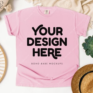 May include: A light pink t-shirt with the text "YOUR DESIGN HERE" in bold black letters. The shirt is a mockup, with rolled-up sleeves. The words "BOHO BABE MOCKUPS" are printed at the bottom of the shirt.