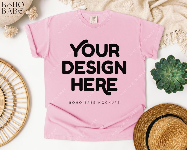 May include: A light pink t-shirt with the text "YOUR DESIGN HERE" in bold black letters. The shirt is a mockup, with rolled-up sleeves. The words "BOHO BABE MOCKUPS" are printed at the bottom of the shirt.