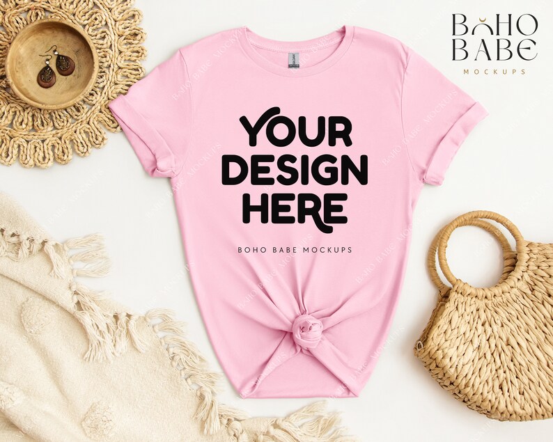 May include: Pink t-shirt mockup with the text "YOUR DESIGN HERE" and the words "BOHO BABE MOCKUPS" below. The t-shirt is laid flat on a white surface with a woven basket and a blanket.