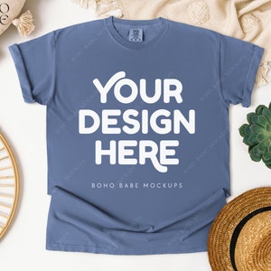 May include: A dusty blue t-shirt with the text "YOUR DESIGN HERE" in white. The shirt is laid flat, with rolled-up sleeves. The image is styled with a straw hat, a succulent, and a woven decorative piece.