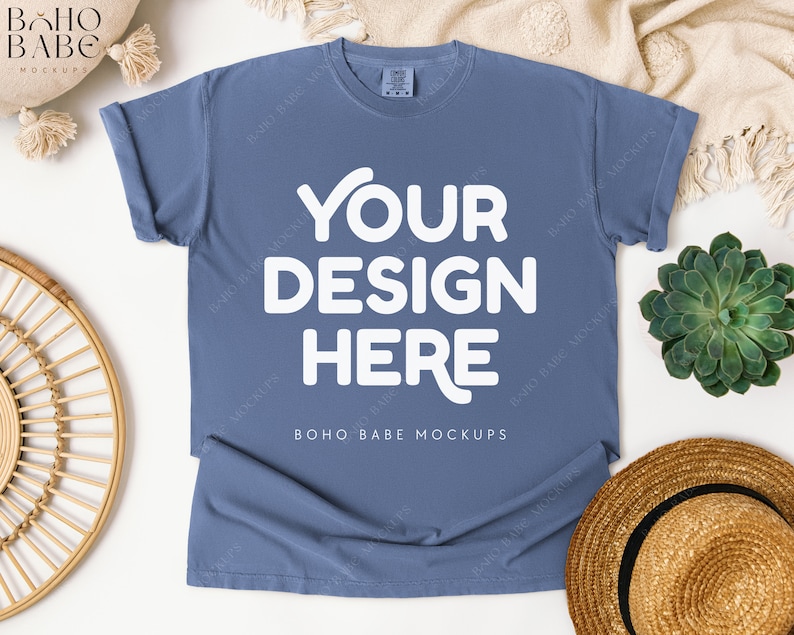 May include: A dusty blue t-shirt with the text "YOUR DESIGN HERE" in white. The shirt is laid flat, with rolled-up sleeves. The image is styled with a straw hat, a succulent, and a woven decorative piece.