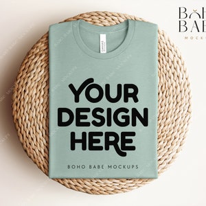 Może przedstawiać: Makieta koszulki w kolorze szałwiowej zieleni na plecionym podkładce ze słomy. Na koszulce znajduje się napis "YOUR DESIGN HERE" wydrukowany czarnym kolorem.