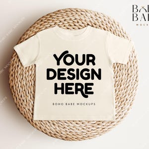 Könnte beinhalten: Ein cremefarbenes T-Shirt mit einem schwarzen Textüberlagerung, auf dem "YOUR DESIGN HERE" und "BOHO BABE MOCKUPS" steht. Das T-Shirt liegt auf einer geflochtenen Strohmatte.