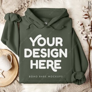 Puede incluir: Una sudadera con capucha verde con el texto "YOUR DESIGN HERE" en blanco en la parte delantera. La sudadera est&aacute; sobre una superficie blanca y peluda con una bandeja de madera y una taza blanca con un dise&ntilde;o de coraz&oacute;n.