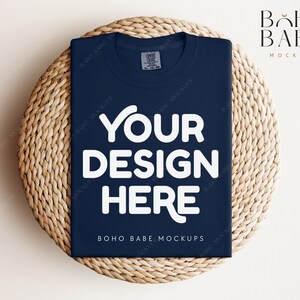 Pode incluir: Uma maquete de camiseta azul marinho com o texto "YOUR DESIGN HERE" em branco sobre um tapete de palha trançado. O texto "BOHO BABE MOCKUPS" também é visível na camiseta.