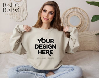 May include: A beige sweatshirt with the text "YOUR DESIGN HERE" and "BOHO BABE MOCKUPS" printed on the front. The model is wearing the sweatshirt with blue jeans and is sitting on a bed with a white blanket.