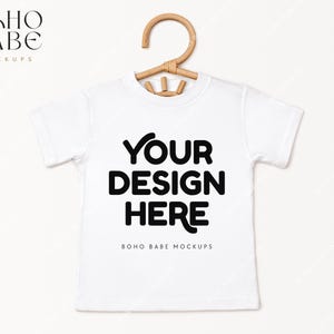 May include: White t-shirt mockup with a wooden hanger. The shirt has the text "YOUR DESIGN HERE" and "BOHO BABE MOCKUPS" printed on it.