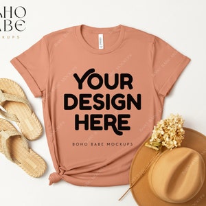 Può includere: Una maglietta color pesca con il testo "YOUR DESIGN HERE" in nero. La maglietta è appoggiata su una superficie bianca con un cappello di paglia e sandali.