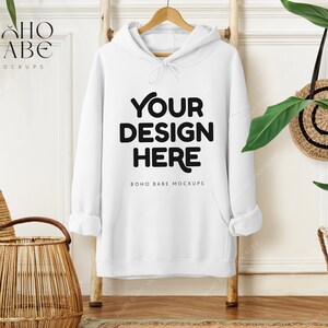 May include: A white hoodie with the text "YOUR DESIGN HERE" in black, hanging on a wooden ladder. The hoodie has a drawstring hood and a front pocket. A woven basket and a round straw bag are also visible.