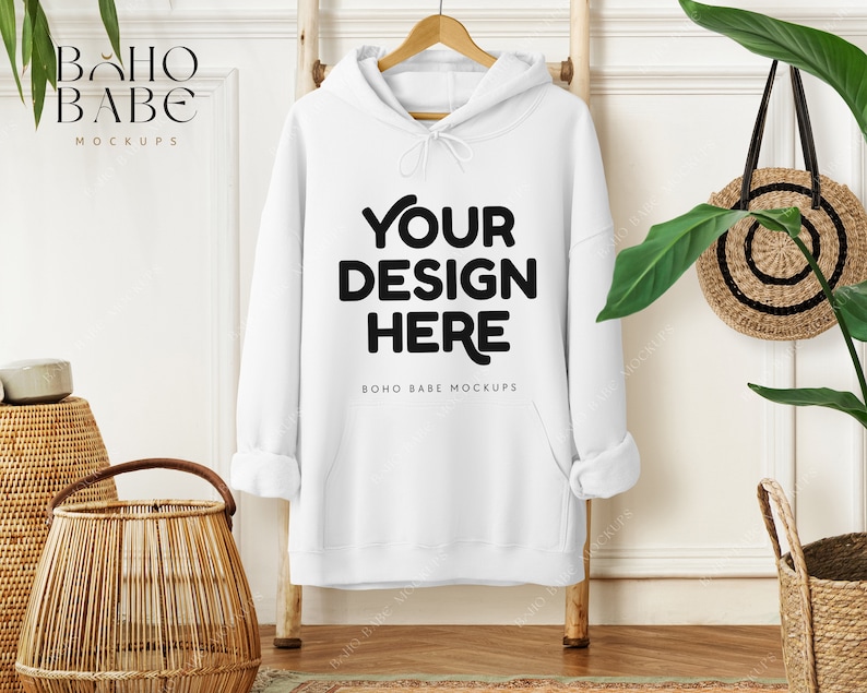 May include: A white hoodie with the text "YOUR DESIGN HERE" in black, hanging on a wooden ladder. The hoodie has a drawstring hood and a front pocket. A woven basket and a round straw bag are also visible.