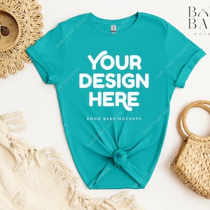 May include: A turquoise blue t-shirt with the text "YOUR DESIGN HERE" in white. The shirt is knotted at the bottom and is laying on a white surface with a woven basket and a white blanket.