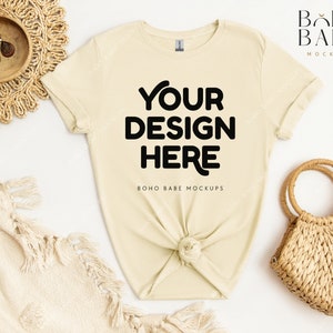 May include: A light beige t-shirt with the text "YOUR DESIGN HERE" in black. The shirt is knotted at the bottom and is laid flat on a white surface. There is a woven straw bag and a woven straw mat in the background.