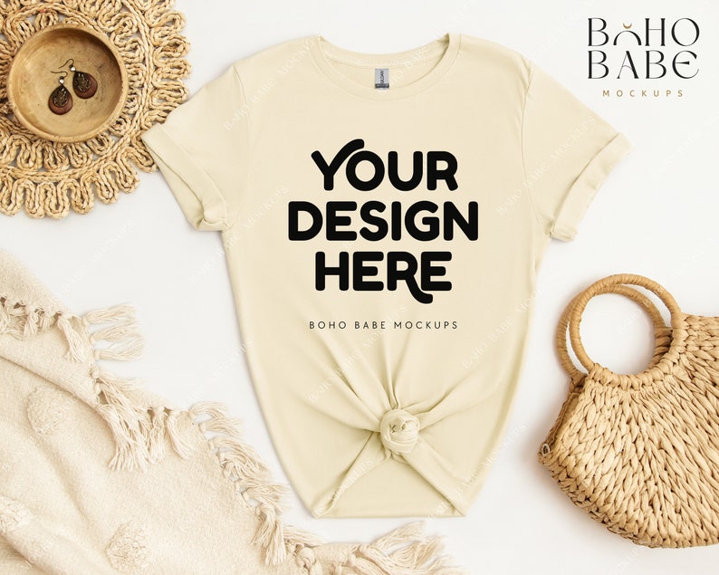 May include: A light beige t-shirt with the text "YOUR DESIGN HERE" in black. The shirt is knotted at the bottom and is laid flat on a white surface. There is a woven straw bag and a woven straw mat in the background.