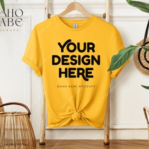 Puede incluir: Una maqueta de camiseta amarilla con el texto "YOUR DESIGN HERE" y "BOHO BABE MOCKUPS" sobre un fondo blanco. La camiseta está colgada de una escalera de madera.