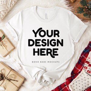 May include: A white t-shirt with the text "YOUR DESIGN HERE" in bold black letters. The shirt is displayed on a white fluffy surface, with wrapped gifts and holiday-themed pajamas. The t-shirt is a mock-up for design placement.