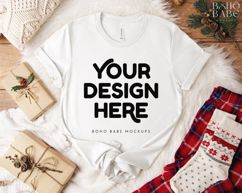 May include: A white t-shirt with the text "YOUR DESIGN HERE" in bold black letters. The shirt is displayed on a white fluffy surface, with wrapped gifts and holiday-themed pajamas. The t-shirt is a mock-up for design placement.