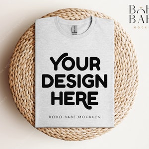 May include: A gray t-shirt with the text "YOUR DESIGN HERE" and "BOHO BABE MOCKUPS" printed on it. The t-shirt is lying flat on a woven straw mat.