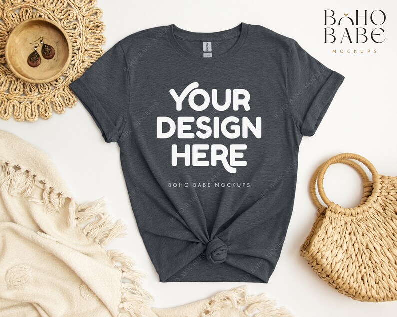 May include: A dark gray t-shirt with the text "YOUR DESIGN HERE" in white. The shirt is knotted at the bottom and is laid out on a white surface with a woven basket and a white blanket.